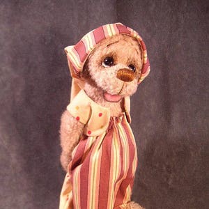 Könnte beinhalten: Ein Plüschteddybär, der ein rosa-gelb gestreiftes Kleid mit passendem Hut trägt. Der Bär hat eine braune Nase und braune Augen. Der Bär hält eine kleine Glocke in seiner Pfote.