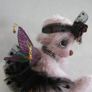 Könnte beinhalten: Ein rosa Mohair-Teddybär, der ein schwarzes Tüll-Tutu, ein schwarzes Stirnband mit schwarz-weißem Tupfenmuster und einen lila Schmetterlingsflügel mit goldenen Akzenten trägt.