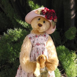 May include: A plush teddy bear wearing a pink hat with red flowers and a floral dress. The bear is holding a smaller plush bear in its arms.