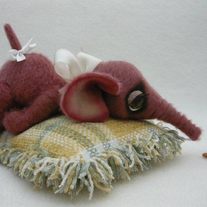 May include: A plush pink elephant toy with a white bow lying on a yellow and green patterned pillow. A small brown toy mouse is standing to the right of the pillow.