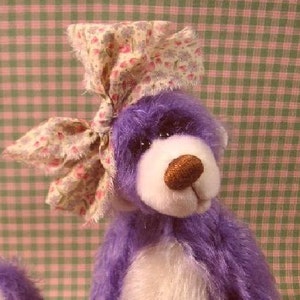 May include: A purple and white teddy bear wearing a floral print fabric bow. The bear has a white face and a brown nose.