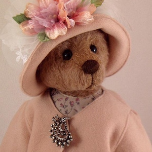 May include: A brown teddy bear wearing a pink hat with a floral trim and a pink coat with a silver pendant.