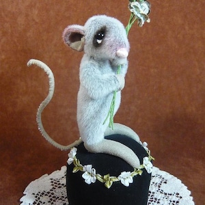 May include: A gray and pink plush mouse holding a bouquet of green and white flowers, sitting on a black velvet pincushion with a white lace trim.