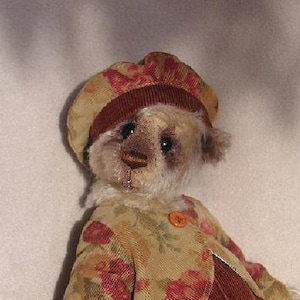 Puede incluir: Un oso de peluche que lleva un gorro de color marrón y beige con estampado y una chaqueta a juego con estampado floral. El oso tiene la cara blanca con ojos marrones y nariz marrón.