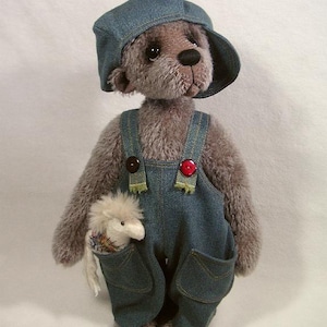 Può includere: Un orsacchiotto di peluche grigio che indossa un cappello e una salopette di jeans blu con bottoni rossi. L'orso tiene un piccolo peluche bianco nella sua zampa destra.