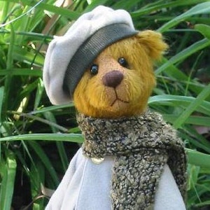May include: A yellow teddy bear wearing a white beret with a black band and a brown and gold scarf. The bear is sitting in front of green foliage.