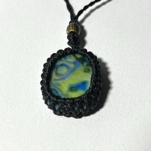 以下が含まれることがあります： 手作りのマクラメネックレス。楕円形のペンダントが付いています。ペンダントは、緑、青、黄色の鮮やかな渦巻き模様です。ペンダントは黒いマクラメ織りで囲まれ、ネックレスは金色の留め金が付いた黒いコードが付いています。