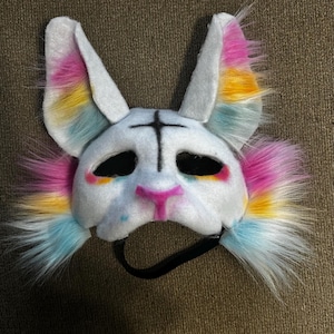 May include: A white animal mask featuring tall ears and fluffy faux fur accents in pink, yellow, and blue. The mask has black eye cutouts, a black cross symbol on the forehead, a pink nose, and a black elastic strap attached to the bottom edge.
