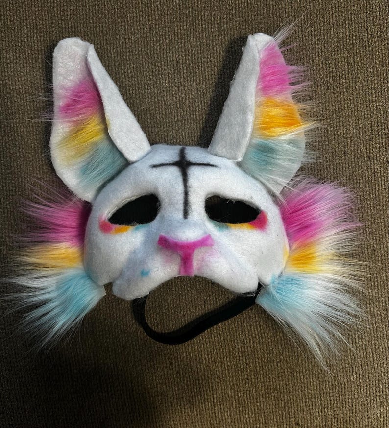 May include: A white animal mask featuring tall ears and fluffy faux fur accents in pink, yellow, and blue. The mask has black eye cutouts, a black cross symbol on the forehead, a pink nose, and a black elastic strap attached to the bottom edge.