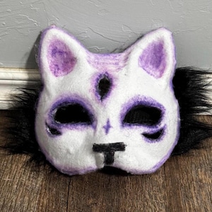 May include: A white cat mask with purple accents around the eyes, ears, and nose. The mask has black eye holes and a black nose. Black faux fur surrounds the mask, giving it a fluffy appearance.