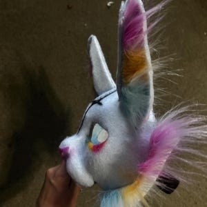 May include: A light blue animal mask featuring tall, pointed ears with multi-coloured faux fur in shades of pink, orange, and blue. The mask has a soft, plush texture, a dark nose, and detailed eye openings with colourful accents around the edges.