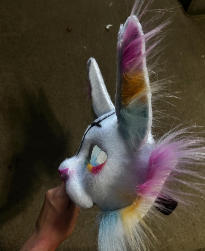 May include: A light blue animal mask featuring tall, pointed ears with multi-coloured faux fur in shades of pink, orange, and blue. The mask has a soft, plush texture, a dark nose, and detailed eye openings with colourful accents around the edges.