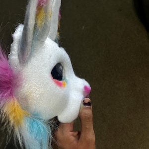 May include: A white plush animal head mask featuring large pointed ears with pink and yellow accents. The mask has a smooth white face, a small pink nose, and a large dark eye. Long, soft faux fur in shades of pink, yellow, and blue extends from the back.