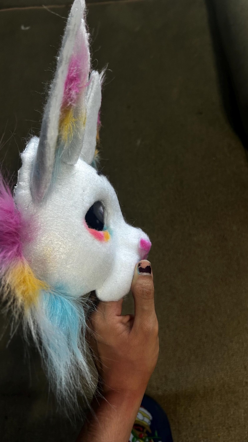 May include: A white plush animal head mask featuring large pointed ears with pink and yellow accents. The mask has a smooth white face, a small pink nose, and a large dark eye. Long, soft faux fur in shades of pink, yellow, and blue extends from the back.