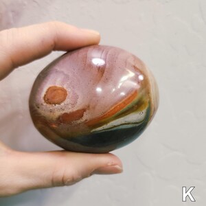 May include: A polished, smooth, oval-shaped stone featuring a multi-coloured surface with bands of mauve, rust-orange, cream, and dark charcoal grey. The stone displays a distinct circular brown inclusion on its left side and a glossy finish.