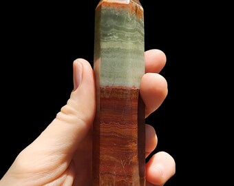 May include: A layered, polished crystal obelisk with a pointed top. The stone features bands of reddish-brown, green, and tan hues. The obelisk is held in a hand against a black background, showcasing its natural colours and textures.