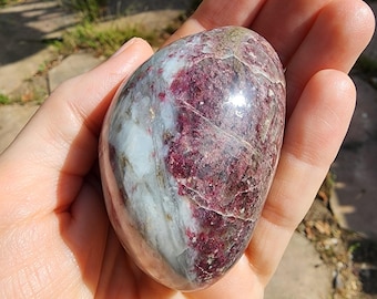 May include: A polished, oval-shaped gemstone with a mix of colors. The stone features a blend of deep burgundy, white, and gray hues, with areas of translucence. The surface is smooth and reflective, suggesting a high polish. The stone is held in a hand.
