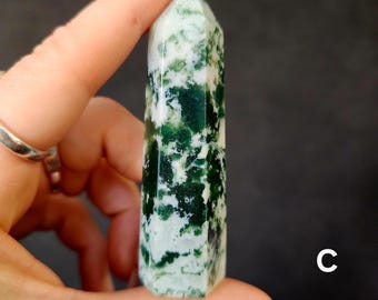 May include: A polished, green and white crystal obelisk is held between fingers against a dark background. The crystal has a pointed top and a pattern of green inclusions within the white stone. The letter "C" is visible in the lower right corner.