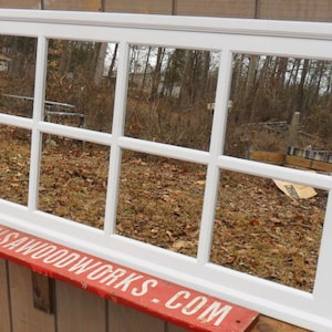 May include: White framed mirror with nine panes, reflecting a view of trees and leaves. The mirror is mounted on a wooden wall.