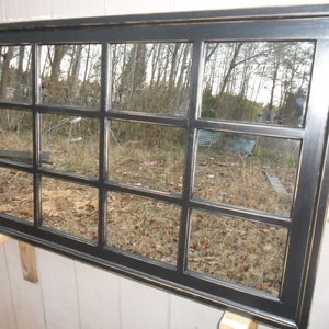May include: A large black framed mirror with a grid of 12 rectangular panes. The mirror reflects a view of trees and foliage.