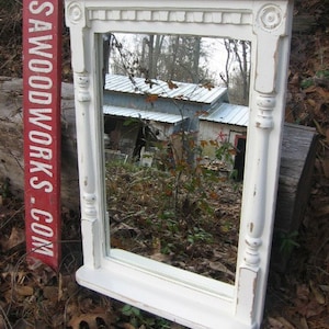 May include: White painted wood framed mirror with a shelf. The mirror has a decorative top with a carved design. The mirror is reflecting a view of a building and trees.