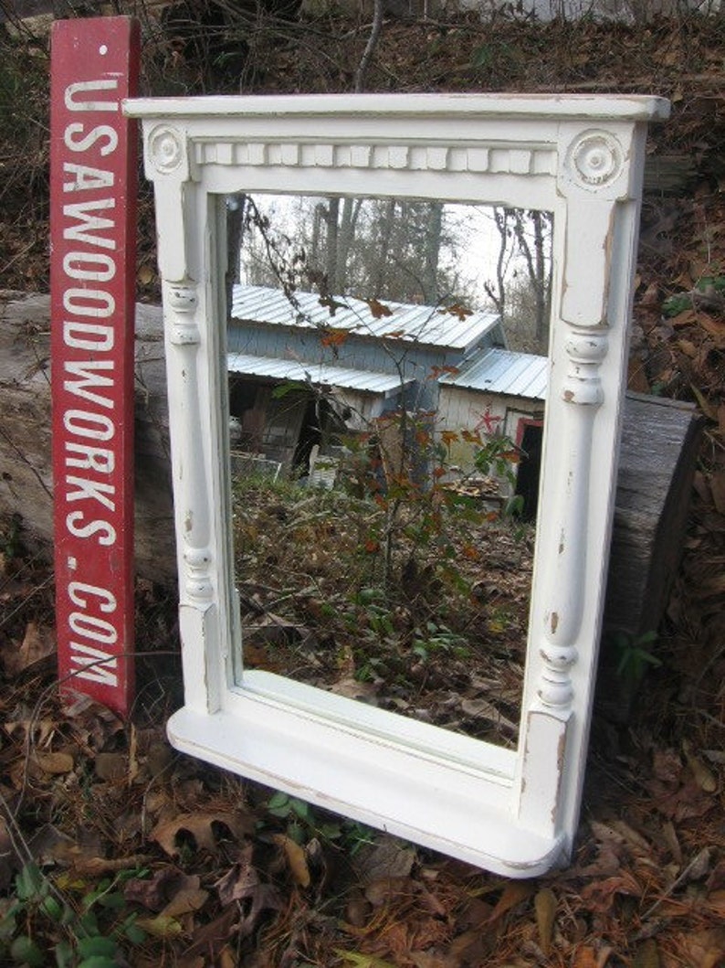 May include: White painted wood framed mirror with a shelf. The mirror has a decorative top with a carved design. The mirror is reflecting a view of a building and trees.