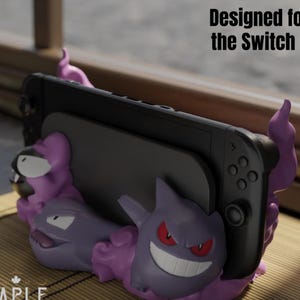 May include: A black Nintendo Switch console is held by a purple and lavender 3D-printed stand featuring two cartoon ghost characters. The stand has the text "Designed for the Switch 2".