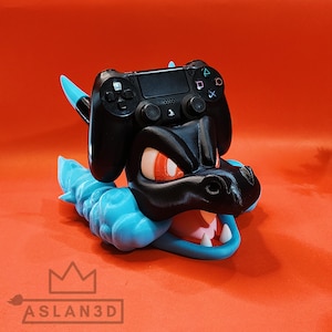Może przedstawiać: Czarny kontroler PlayStation 4 spoczywa na drukowanym w 3D uchwycie na kontroler w stylu Charizarda. Uchwyt jest czarny z pomarańczowymi oczami, białymi zębami i niebieskimi akcentami. Na podstawie znajduje się napis "ASLAN3D".