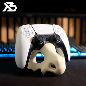 May include: A white and black PlayStation 5 controller with a bone-colored skull-shaped stand. The controller has black joysticks and buttons, and the stand is designed to hold the controller upright. The logo "B" is in the top left corner.