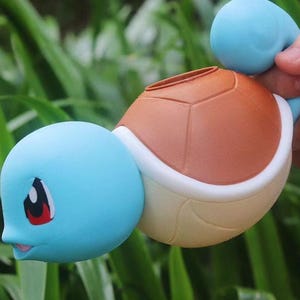 以下が含まれることがあります： 青、茶色、白のゼニガメのフィギュア。フィギュアは、水色の頭と尾、茶色の甲羅、白いお腹をしています。ゼニガメは赤と黒の目と笑顔の口をしています。フィギュアは手で持たれています。