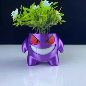 Puede incluir: Una maceta morada con forma de personaje de dibujos animados, con ojos rojos y una amplia sonrisa, que contiene follaje verde y flores azul claro. La maceta tiene brazos y piernas pequeños y está sobre una superficie blanca.