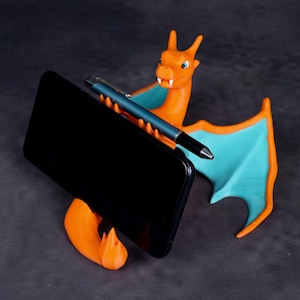 May include: An orange and teal dragon-shaped phone stand holding a black smartphone and a blue pen. The dragon has outstretched wings and a flame-like base, perfect for a desk or table. The stand is made of plastic.