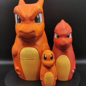 May include: Three orange figurines of the popular Charmander evolution line from the Pokémon franchise. The largest figure is Charizard, the middle is Charmeleon, and the smallest is Charmander. Each figure has a yellow belly and white accents.