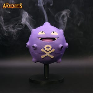May include: A purple, cartoon-like figure with a skull and crossbones symbol, mounted on a black stand. The figure has a smiling face and is emitting white smoke. The Alchemists logo is in the top left corner.