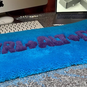 Puede incluir: Una alfombra rectangular azul brillante con las palabras "CTRL+ALT+DEL" en letras moradas y esponjosas. La alfombra está colocada sobre una superficie con un teclado, una computadora y un ratón en el fondo.