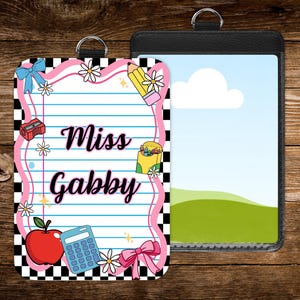 May include: Two ID badge holders. One features a pink and black checkered border with school-themed illustrations, including an apple, pencil, and calculator, and the text "Miss Gabby." The other is a black holder with a landscape scene.