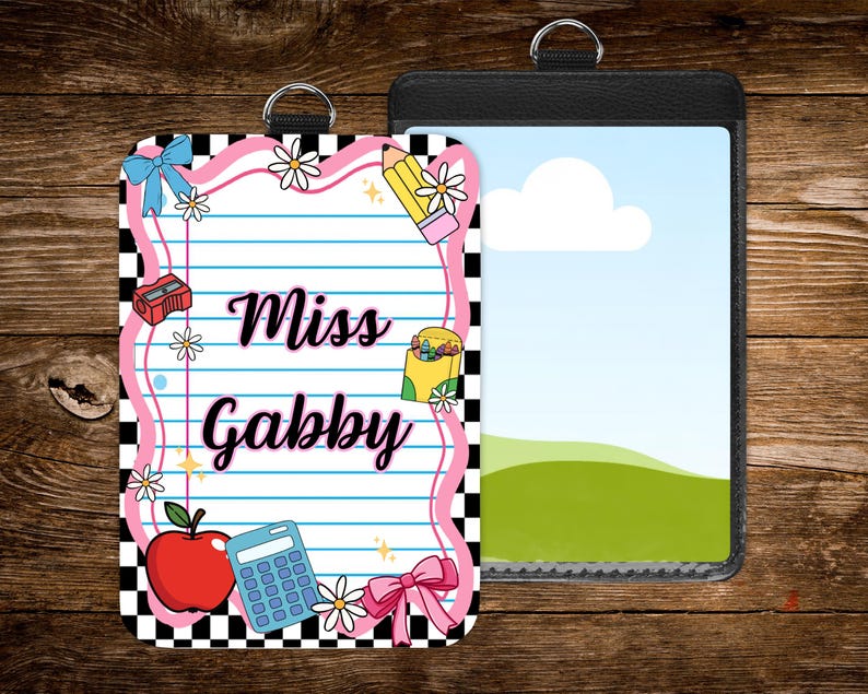 May include: Two ID badge holders. One features a pink and black checkered border with school-themed illustrations, including an apple, pencil, and calculator, and the text "Miss Gabby." The other is a black holder with a landscape scene.