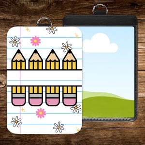 May include: Two ID badge holders. One features a white background with a pencil and eraser design, blue lines, and flower accents. The other is a black leather-like holder with a landscape image. Both have a silver ring for attachment.