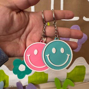 May include: Two round keyrings with smiley faces. One is pink, the other is teal. Both have white smiles and eyes. The keyrings are held up by a silver chain and ring. The background has floral and bunny decorations.