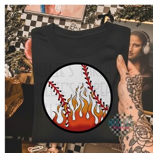 May include: Black t-shirt featuring a baseball graphic with red stitching and flames in shades of orange and yellow. The baseball design is centered on the shirt. The shirt is folded and held by a person.