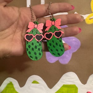 Pode incluir: Dois chaveiros verdes em forma de picles com detalhes de pontos pretos, cada um adornado com óculos de sol rosa em forma de coração e um laço rosa. Os chaveiros penduram de correntes de bolinhas prateadas. O fundo é de cor castanho claro.