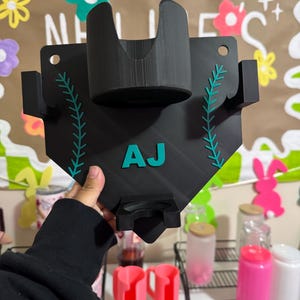 May include: Black baseball-shaped holder with teal accents and the letters "AJ". The holder has a cup holder and a larger curved section. Teal stitching-like design on the sides. Additional pink cup holders are visible.