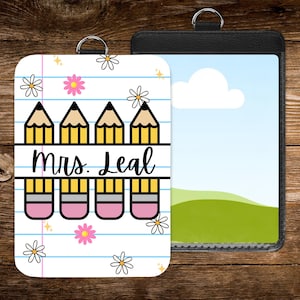 May include: A white and pink teacher badge holder with a pencil and eraser design, the text "Mrs. Leal", and daisy accents. A black badge holder with a landscape image is next to it. Both have silver rings.