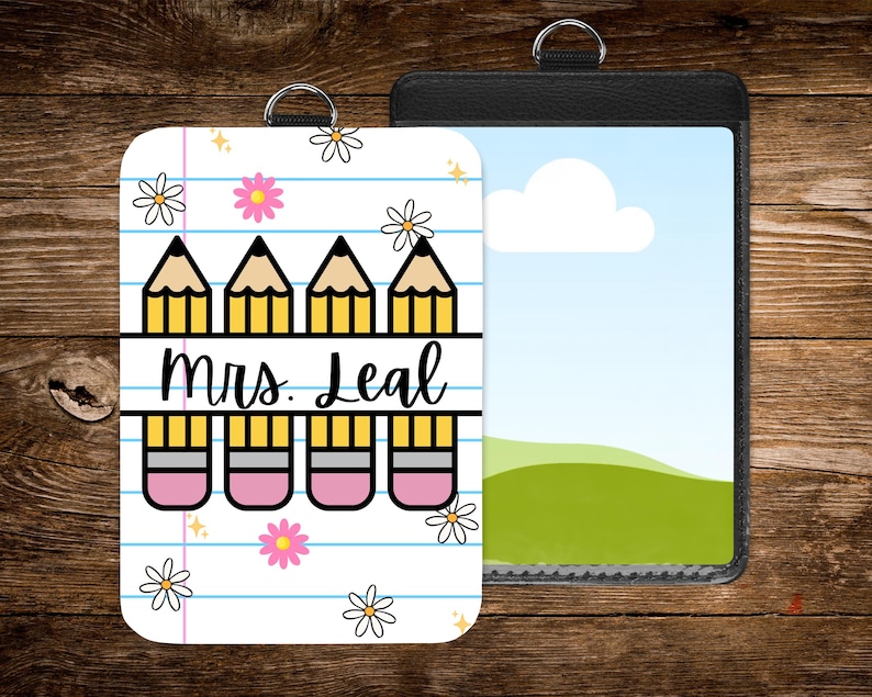May include: A white and pink teacher badge holder with a pencil and eraser design, the text "Mrs. Leal", and daisy accents. A black badge holder with a landscape image is next to it. Both have silver rings.