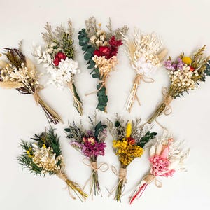 Puede incluir: Vista cenital de nueve peque&ntilde;os ramos de flores secas. Cada ramo, atado con hilo, presenta una variedad de colores, incluyendo blanco, rojo, amarillo y morado. Los ramos est&aacute;n dispuestos sobre una superficie blanca.