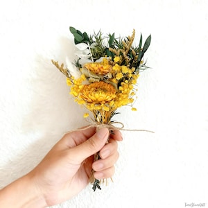 Puede incluir: Un peque&ntilde;o ramo de flores secas con flores amarillas brillantes y acentos verdes y blancos. El ramo est&aacute; atado con hilo y sostenido en una mano sobre un fondo blanco. Las flores tienen varias texturas y formas.