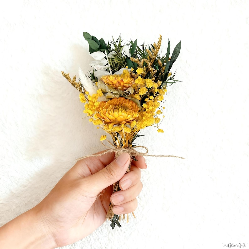 Puede incluir: Un peque&ntilde;o ramo de flores secas con flores amarillas brillantes y acentos verdes y blancos. El ramo est&aacute; atado con hilo y sostenido en una mano sobre un fondo blanco. Las flores tienen varias texturas y formas.