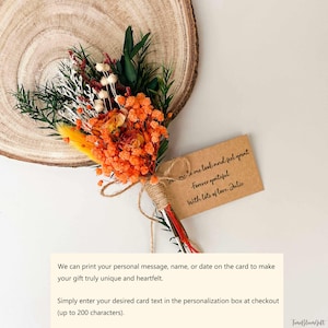 Puede incluir: Un peque&ntilde;o ramo de flores secas con elementos naranjas, amarillos, blancos y verdes, atado con hilo. Se adjunta una peque&ntilde;a tarjeta con texto escrito a mano. El ramo descansa sobre una rodaja de madera.