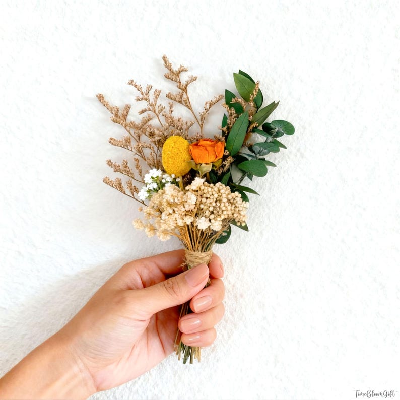 Puede incluir: Un peque&ntilde;o ramo de flores secas, que incluye flores amarillas, naranjas y blancas, y hojas verdes, atadas con hilo. El ramo se sostiene en una mano sobre un fondo blanco.