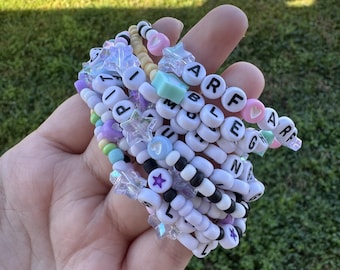 Puede incluir: Una pila de pulseras de cuentas de colores con abalorios de letras y estrellas. Las pulseras están hechas con cuentas blancas, negras, rosas, azul claro y verde claro.